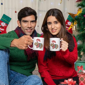 Pu&ograve; includere: Due persone tengono in mano tazze bianche con un design di capibara natalizio. Le tazze presentano un capibara con un maglione festivo e un cappello da Babbo Natale. Lo sfondo include un albero di Natale, calze e regali confezionati, creando un tema festivo.