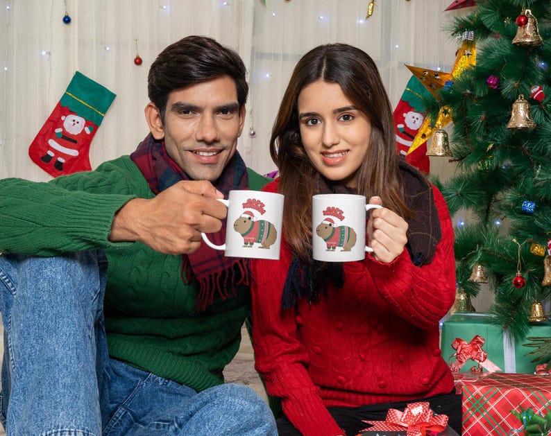 Pu&ograve; includere: Due persone tengono in mano tazze bianche con un design di capibara natalizio. Le tazze presentano un capibara con un maglione festivo e un cappello da Babbo Natale. Lo sfondo include un albero di Natale, calze e regali confezionati, creando un tema festivo.