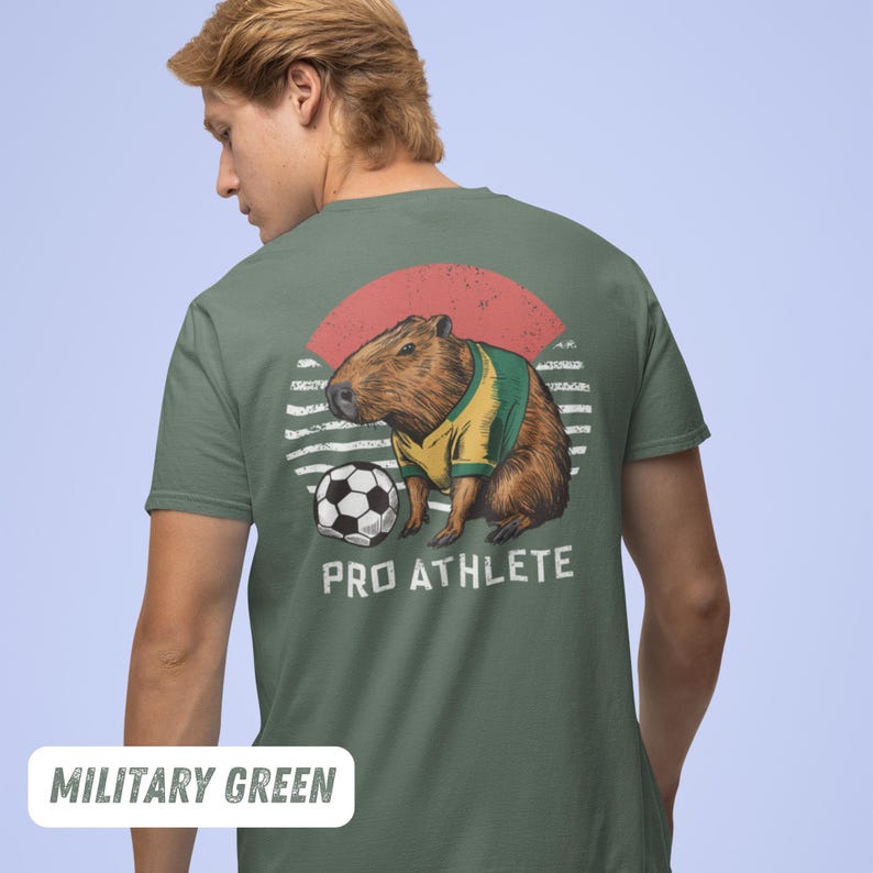 Puede incluir: Una camiseta verde militar con un gr&aacute;fico de una capibara vistiendo una camiseta de f&uacute;tbol y sosteniendo un bal&oacute;n de f&uacute;tbol. El gr&aacute;fico est&aacute; rodeado por un sol radiante rojo, blanco y amarillo. El texto "PRO ATHLETE" est&aacute; impreso debajo del gr&aacute;fico.
