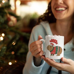 Pu&ograve; includere: Tazza in ceramica bianca con un capibara che indossa un maglione natalizio e un cappello da Babbo Natale. La tazza presenta la scritta "Merry Christmas" sopra il capibara. Lo sfondo include un albero di Natale con luci.