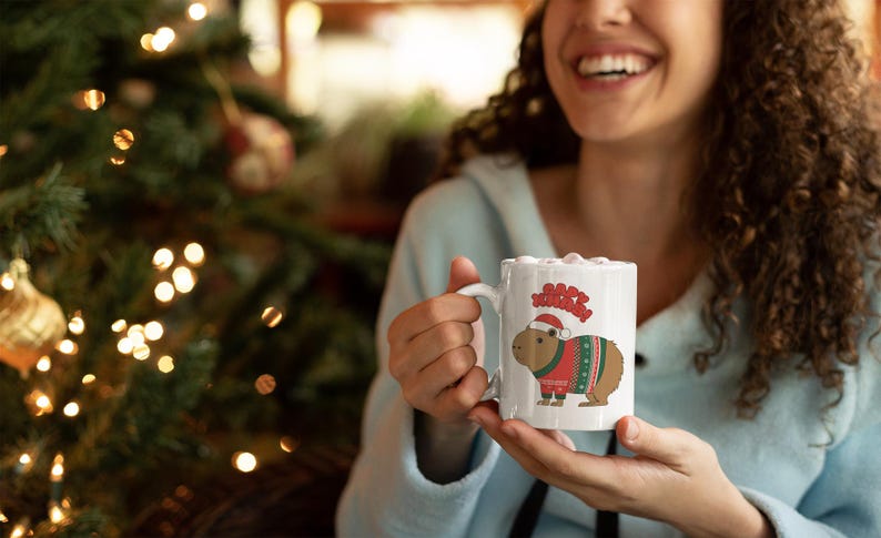 Pu&ograve; includere: Tazza in ceramica bianca con un capibara che indossa un maglione natalizio e un cappello da Babbo Natale. La tazza presenta la scritta "Merry Christmas" sopra il capibara. Lo sfondo include un albero di Natale con luci.