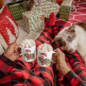 Pu&ograve; includere: Due tazze bianche piene di marshmallow, ognuna con un capibara con un maglione natalizio e un cappello da Babbo Natale. La persona indossa un pigiama a quadri rossi e neri. Un cane guarda la fotocamera. Albero di Natale e regali incartati sullo sfondo.