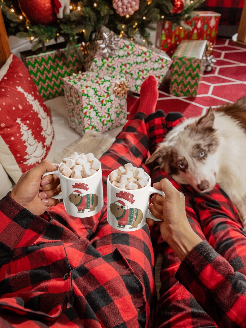 Pu&ograve; includere: Due tazze bianche piene di marshmallow, ognuna con un capibara con un maglione natalizio e un cappello da Babbo Natale. La persona indossa un pigiama a quadri rossi e neri. Un cane guarda la fotocamera. Albero di Natale e regali incartati sullo sfondo.
