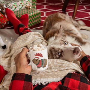 Pu&ograve; includere: Una tazza di ceramica bianca piena di marshmallow, con un capibara dei cartoni animati con un cappello di Babbo Natale e un maglione natalizio. La tazza &egrave; tenuta da una persona che indossa una camicia a quadri rossa e nera, con un cane e regali di Natale sullo sfondo.