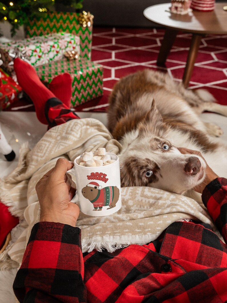 Pu&ograve; includere: Una tazza di ceramica bianca piena di marshmallow, con un capibara dei cartoni animati con un cappello di Babbo Natale e un maglione natalizio. La tazza &egrave; tenuta da una persona che indossa una camicia a quadri rossa e nera, con un cane e regali di Natale sullo sfondo.