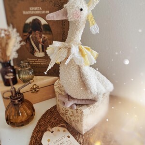 May include: A plush white goose toy with a pink beak and a yellow and white ruffled collar. The goose sits atop a decorative box with a floral pattern. A tag with the text "MOONY ZOO" is visible.