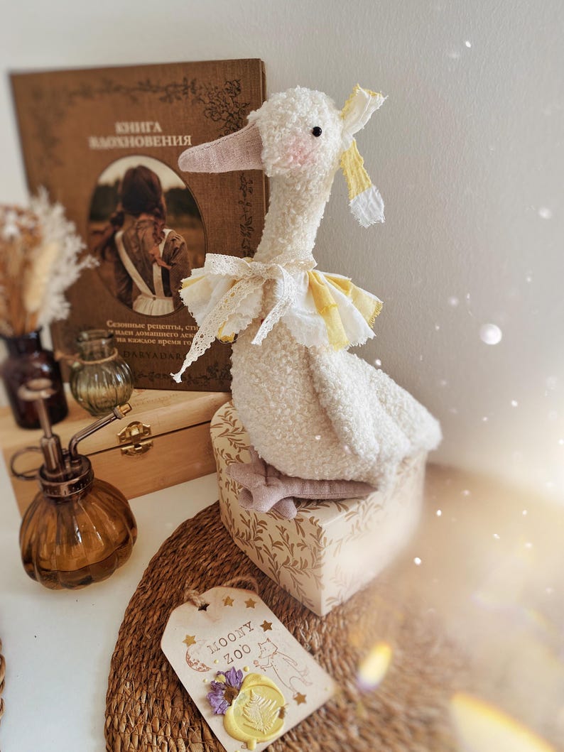 May include: A plush white goose toy with a pink beak and a yellow and white ruffled collar. The goose sits atop a decorative box with a floral pattern. A tag with the text "MOONY ZOO" is visible.