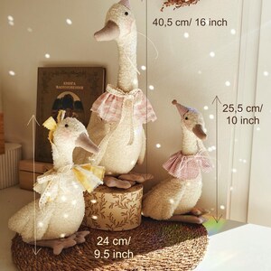 May include: Three plush goose dolls of varying sizes. The tallest is 16 inches, the middle one is 9.5 inches, and the smallest is 10 inches. Each goose wears a hat and a decorative collar. The dolls are cream-colored with pink and yellow accents.