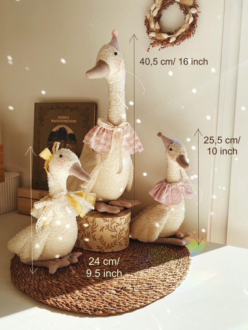 May include: Three plush goose dolls of varying sizes. The tallest is 16 inches, the middle one is 9.5 inches, and the smallest is 10 inches. Each goose wears a hat and a decorative collar. The dolls are cream-colored with pink and yellow accents.