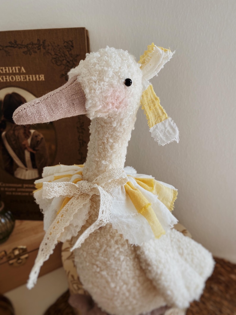 May include: A handmade plush goose doll with a white, textured body, a pink beak, and a black eye. The goose wears a yellow and white ruffled collar and a matching bow. A vintage book is in the background.