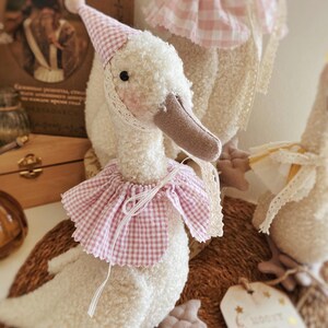 May include: A plush white goose toy with a pink and white checkered party hat and collar. The goose has a brown beak and black eyes. The toy is decorated with lace trim and is part of a collection of similar goose toys.