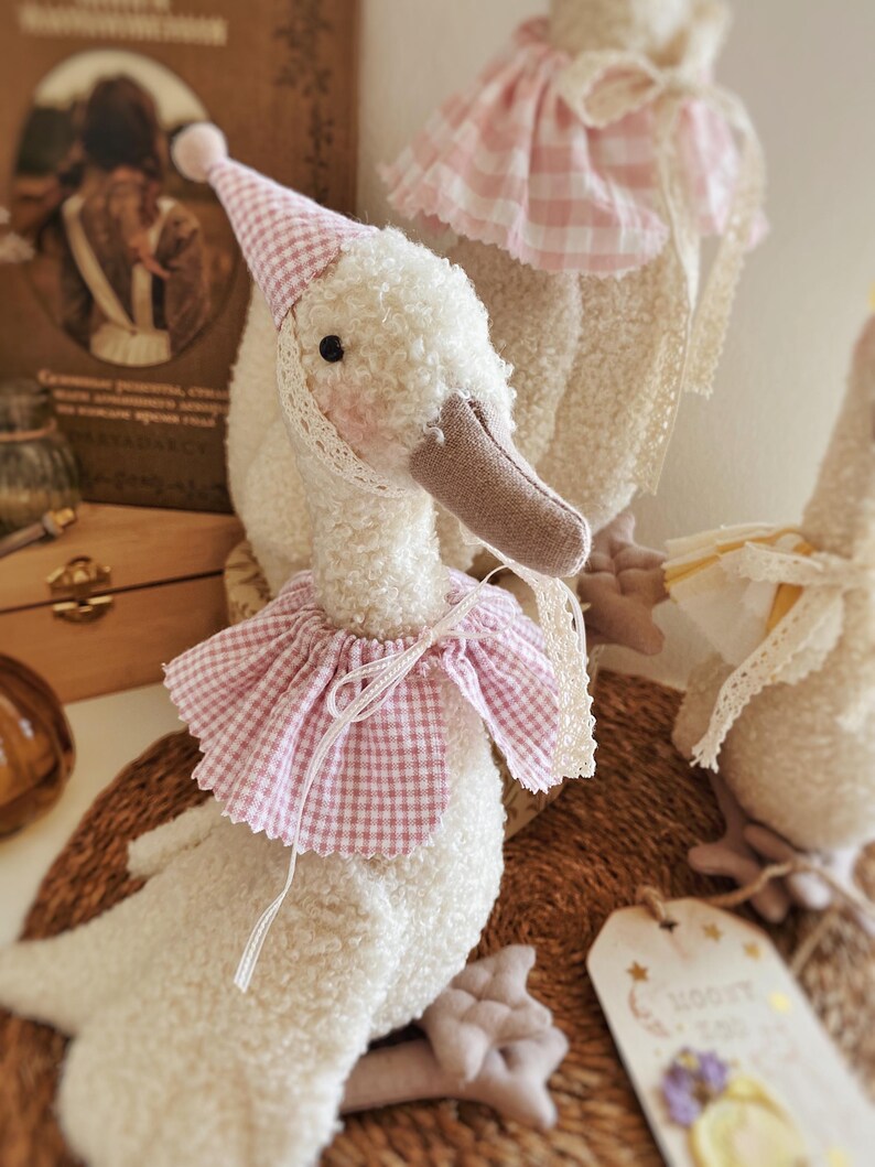 May include: A plush white goose toy with a pink and white checkered party hat and collar. The goose has a brown beak and black eyes. The toy is decorated with lace trim and is part of a collection of similar goose toys.