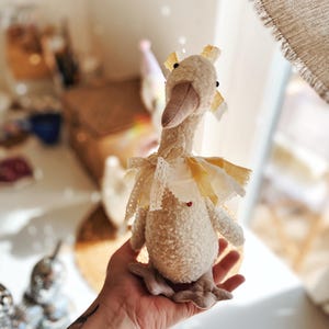 May include: A plush goose doll with a cream-colored body, a beige beak, and a yellow and white checkered collar. The goose has a red heart detail and is adorned with lace and ribbon. The doll is a soft toy.