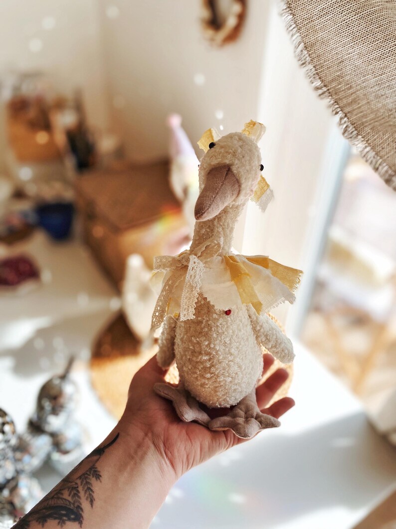 May include: A plush goose doll with a cream-colored body, a beige beak, and a yellow and white checkered collar. The goose has a red heart detail and is adorned with lace and ribbon. The doll is a soft toy.