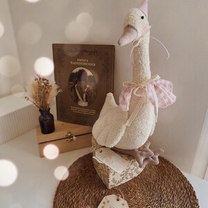 May include: A plush white goose toy with a pink party hat and a pink and white gingham collar. The goose is perched on a decorative box with a floral design. A book with Russian text and a small vase with dried flowers are in the background.