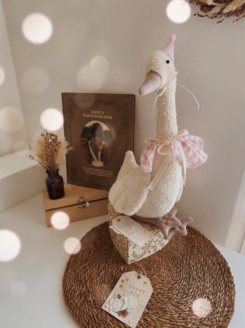 May include: A plush white goose toy with a pink party hat and a pink and white gingham collar. The goose is perched on a decorative box with a floral design. A book with Russian text and a small vase with dried flowers are in the background.