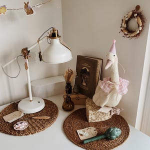 May include: A white desk with a white lamp, a stuffed goose wearing a pink party hat, and a decorative wreath. The desk also features a woven placemat, a small dish, and a decorative artichoke. A string of Easter-themed decorations hangs above.