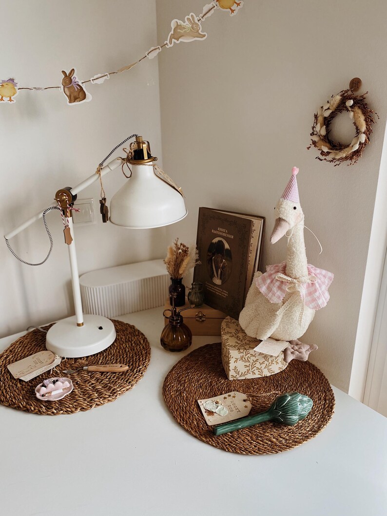 May include: A white desk with a white lamp, a stuffed goose wearing a pink party hat, and a decorative wreath. The desk also features a woven placemat, a small dish, and a decorative artichoke. A string of Easter-themed decorations hangs above.
