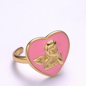 Puede incluir: Anillo dorado con una cara en forma de coraz&oacute;n rosa y un dise&ntilde;o de querub&iacute;n dorado. El anillo tiene un acabado liso y pulido y una banda sencilla. El querub&iacute;n est&aacute; detallado con alas y una pose pensativa.