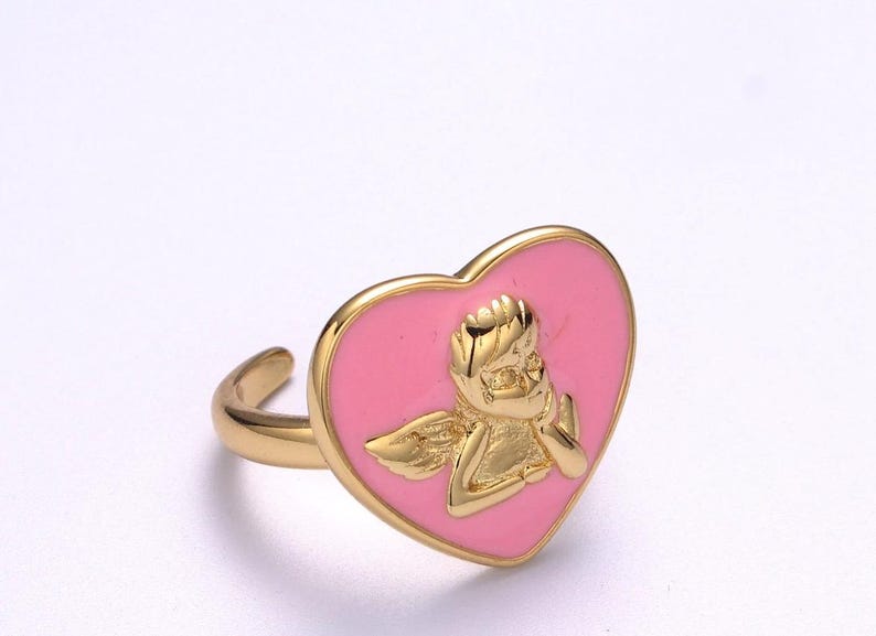Puede incluir: Anillo dorado con una cara en forma de coraz&oacute;n rosa y un dise&ntilde;o de querub&iacute;n dorado. El anillo tiene un acabado liso y pulido y una banda sencilla. El querub&iacute;n est&aacute; detallado con alas y una pose pensativa.