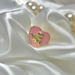 Puede incluir: Un anillo dorado con una cara rosa en forma de coraz&oacute;n con un dise&ntilde;o de querub&iacute;n dorado. El anillo descansa sobre una tela de sat&eacute;n blanco, con un collar de perlas en el fondo.