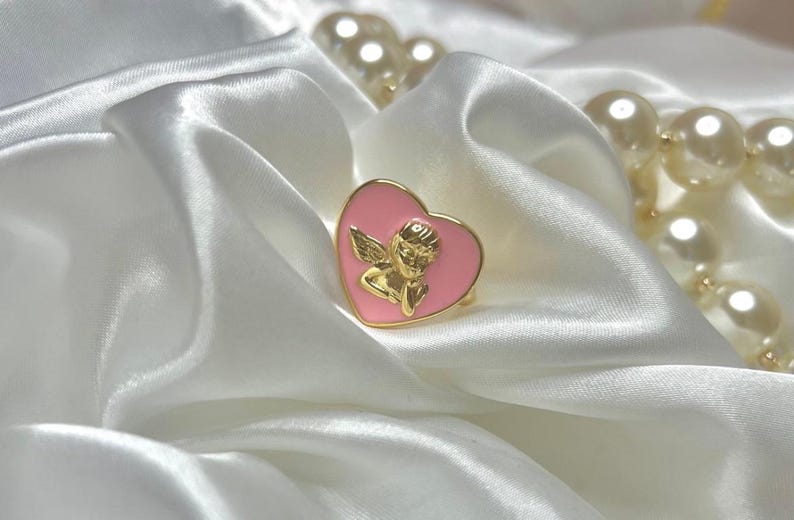Puede incluir: Un anillo dorado con una cara rosa en forma de coraz&oacute;n con un dise&ntilde;o de querub&iacute;n dorado. El anillo descansa sobre una tela de sat&eacute;n blanco, con un collar de perlas en el fondo.