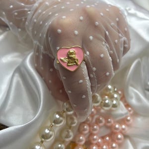 Puede incluir: Un anillo dorado con una cara en forma de coraz&oacute;n rosa con un dise&ntilde;o de querub&iacute;n dorado. El anillo se encuentra sobre un fondo de sat&eacute;n blanco y una mano con un guante blanco con lunares. Tambi&eacute;n se ven collares de perlas blancas y rosas.