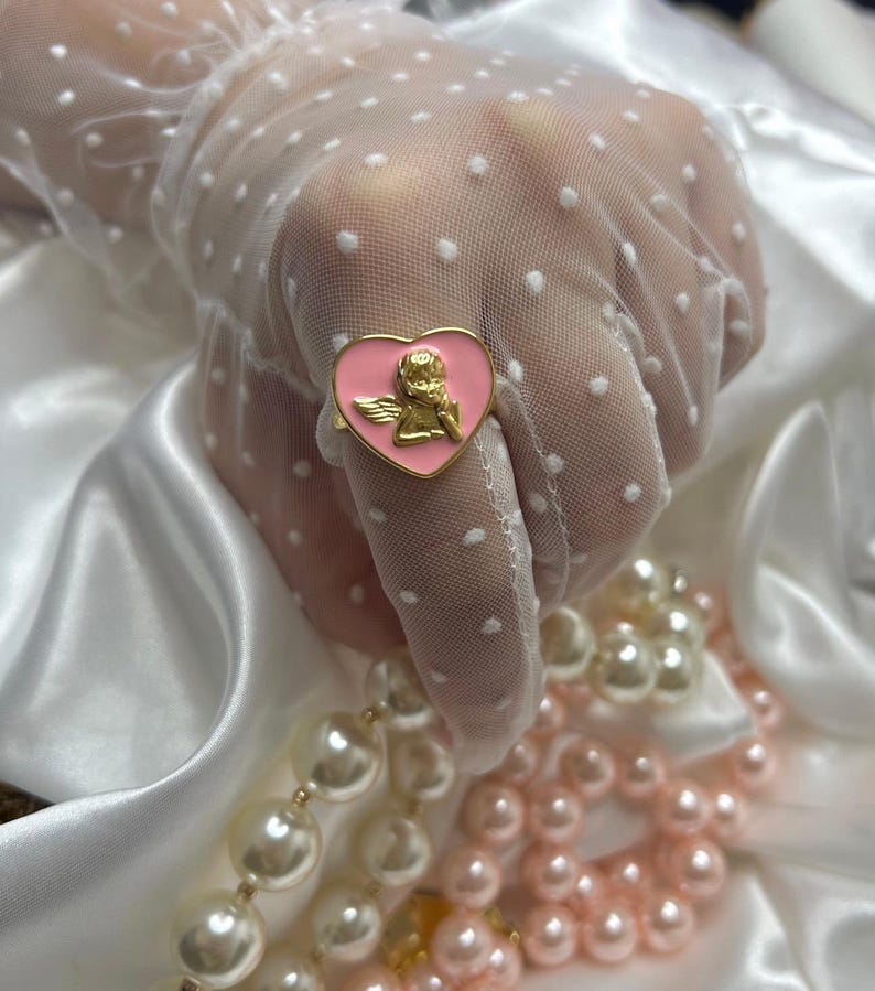 Puede incluir: Un anillo dorado con una cara en forma de coraz&oacute;n rosa con un dise&ntilde;o de querub&iacute;n dorado. El anillo se encuentra sobre un fondo de sat&eacute;n blanco y una mano con un guante blanco con lunares. Tambi&eacute;n se ven collares de perlas blancas y rosas.