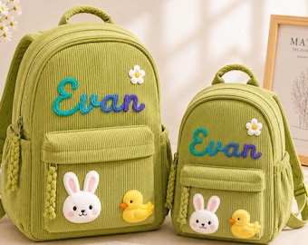 May include: Two olive green corduroy backpacks of different sizes, both featuring the name Evan embroidered in blue and purple, a white flower applique, and two 3D-style character patches of a white rabbit and a yellow duck on the front pocket.