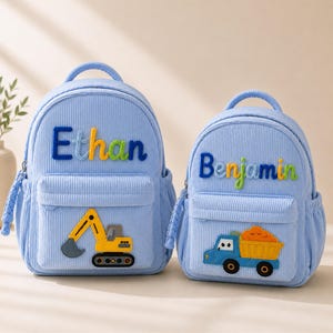 Pode incluir: Duas mochilas de veludo cotelê azul claro com alças superiores e bolsos frontais com zíper. A bolsa maior exibe o nome Ethan em letras bordadas coloridas e uma escavadeira amarela. A menor mostra o nome Benjamin e um caminhão basculante azul.