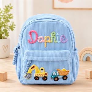 May include: A light blue corduroy backpack featuring the embroidered name "Daphie" in multicolored yarn across the top. The front pocket displays embroidered patches of a yellow construction excavator and a blue and yellow dump truck.