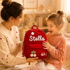 Puede incluir: Una mochila roja con el nombre "Stella" bordado en blanco. La mochila presenta una flor blanca, cerezas y un aplique de zorro. La bolsa es sostenida por una persona y un ni&ntilde;o.