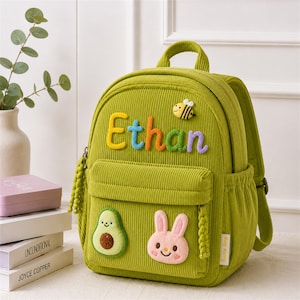 Peut inclure: Un sac à dos en velours côtelé vert lime avec le prénom Ethan en lettres de tissu colorées et en relief. Le sac présente un écusson abeille, un écusson avocat et un écusson tête de lapin rose sur la poche avant, avec des tirettes tressées.