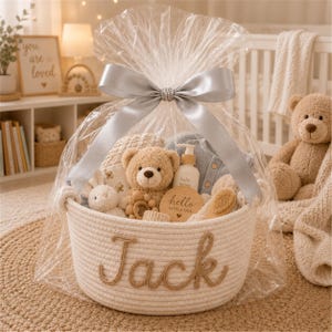 May include: A white rope basket with the name Jack stitched in brown yarn on the front. The basket is wrapped in clear cellophane with a silver ribbon and contains a teddy bear, a wooden rattle, a baby lotion bottle, and a wooden hello little one sign.