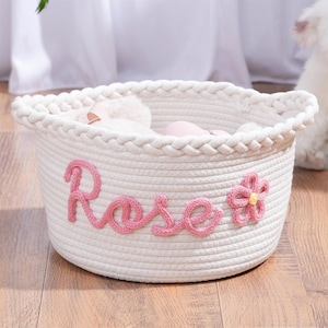 May include: A round white rope storage basket featuring a braided top rim and pink yarn lettering that spells out the name Rose. The basket is decorated with a pink yarn flower appliqu&eacute; with a small yellow centre, constructed from coiled cotton rope.