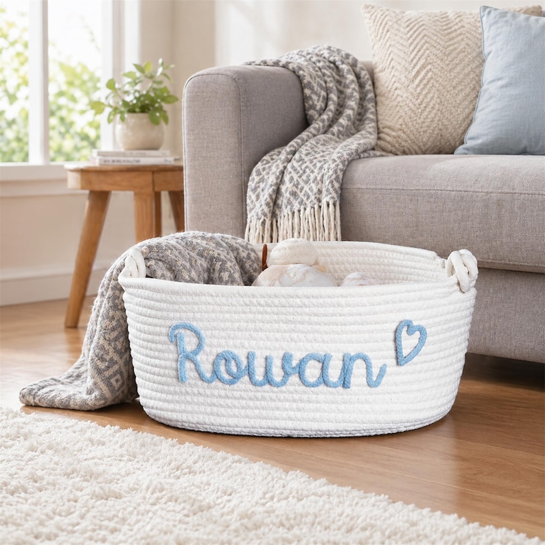 May include: A white oval rope basket featuring the name Rowan embroidered in light blue script lettering with a matching light blue heart icon. The basket has integrated side handles and contains a soft grey patterned textile item inside.