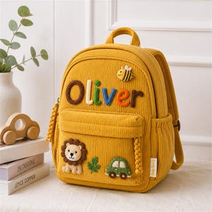Puede incluir: Mochila de pana amarillo mostaza con el nombre "Oliver" en letras coloridas. La mochila presenta apliques de le&oacute;n, abeja, coche y hoja. Tiene un asa superior y bolsillos laterales.