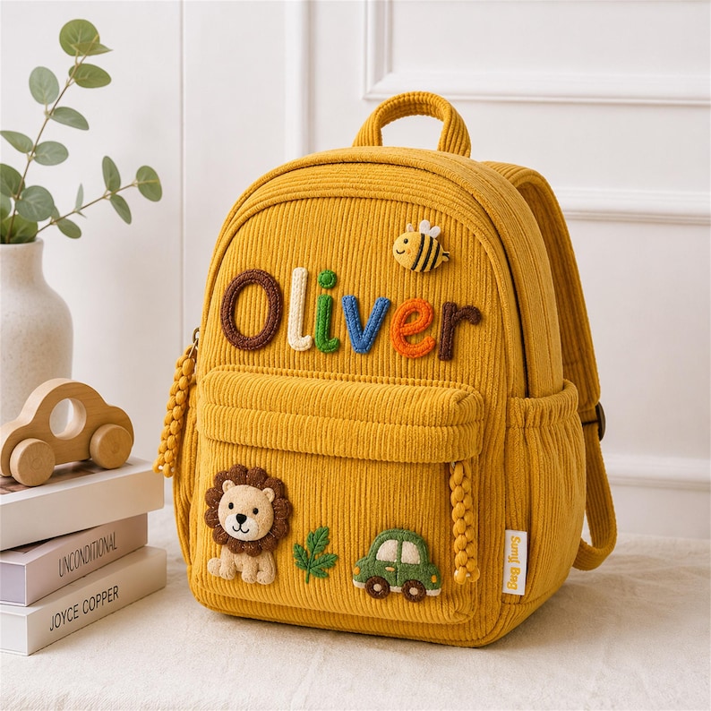 Puede incluir: Mochila de pana amarillo mostaza con el nombre "Oliver" en letras coloridas. La mochila presenta apliques de le&oacute;n, abeja, coche y hoja. Tiene un asa superior y bolsillos laterales.