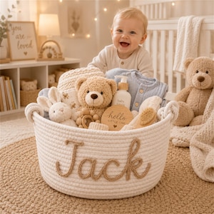 May include: A white woven rope basket featuring the name Jack in brown yarn lettering. The basket contains a plush teddy bear, a white stuffed bunny, a wooden rattle, a bottle of baby lotion, a wooden disc with the text hello LITTLE ONE, and clothing.