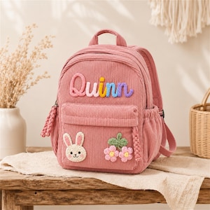 Puede incluir: Mochila de pana rosa con el nombre "Quinn" en letras coloridas. La mochila presenta un bolsillo frontal, un aplique de conejito y detalles florales. Tiene un asa superior y correas ajustables.