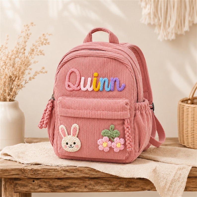 Puede incluir: Mochila de pana rosa con el nombre "Quinn" en letras coloridas. La mochila presenta un bolsillo frontal, un aplique de conejito y detalles florales. Tiene un asa superior y correas ajustables.