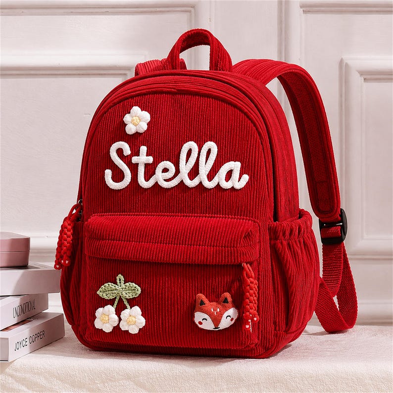 Puede incluir: Mochila de pana roja con el nombre "Stella" en escritura blanca. Incluye un bolsillo frontal, un bolsillo lateral y una peque&ntilde;a aplicaci&oacute;n de zorro. Decorada con una flor y una aplicaci&oacute;n de flor de cerezo. Correas ajustables y un asa superior.