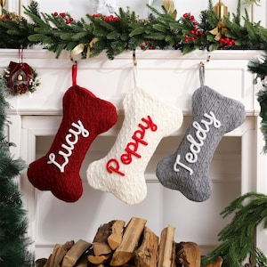 May include: Three bone-shaped Christmas stockings made of cable-knit fabric. The left stocking is dark red with white embroidered text reading Lucy. The center stocking is cream with red text reading Poppy. The right stocking is gray with white text reading Teddy.
