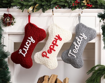 May include: Three bone-shaped Christmas stockings made of cable-knit fabric. The left stocking is dark red with white embroidered text reading Lucy. The center stocking is cream with red text reading Poppy. The right stocking is gray with white text reading Teddy.
