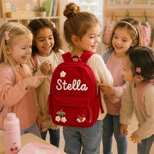 Puede incluir: Un grupo de ni&ntilde;os rodea una mochila roja con el nombre "Stella". La mochila tiene aplicaciones de flores y un zorro. Una botella de agua rosa y una lonchera est&aacute;n sobre una mesa. La escena tiene lugar en un aula.