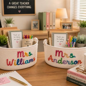 May include: Two oval white cotton rope baskets with integrated handles, each featuring colorful embroidered names, Mrs. Williams and Mrs. Anderson. The baskets contain notebooks labeled Thank you for helping me grow, mugs, and small potted green plants.