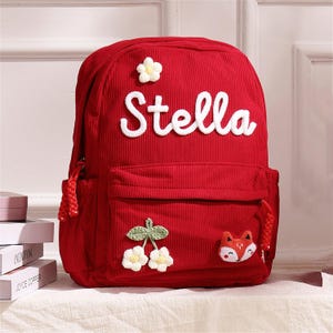 May include: Red corduroy backpack with the name "Stella" in white script. The backpack features a front pocket, a flower and cherry applique, and a fox applique. The backpack has a braided tassel on the side.