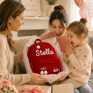 Puede incluir: Una mochila roja con el nombre "Stella" bordado en blanco, decorada con aplicaciones florales y de zorro. La mochila se presenta como un regalo, con un regalo envuelto y una tarjeta que dice "Para ti".
