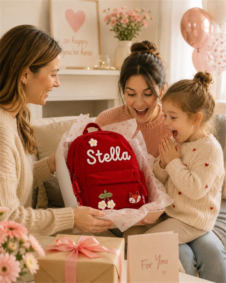 Puede incluir: Una mochila roja con el nombre "Stella" bordado en blanco, decorada con aplicaciones florales y de zorro. La mochila se presenta como un regalo, con un regalo envuelto y una tarjeta que dice "Para ti".