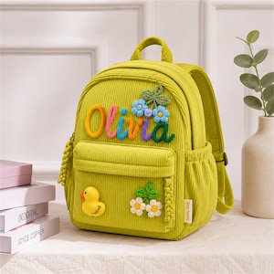 Puede incluir: Una mochila de pana verde lima con el nombre "Olivia" en letras coloridas. La mochila presenta un bolsillo frontal, una aplicaci&oacute;n de pato amarillo y adornos florales. La bolsa tiene correas ajustables y un asa superior.